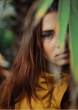 Willow, long auburn hair, emerald green eyes, relaxed facial expression, in yellow rain jacket behind out of focus leaves.